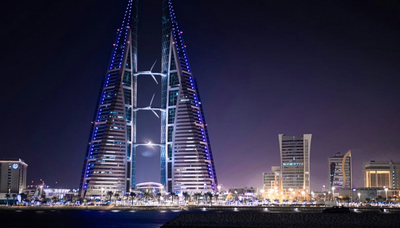 Night view of the illuminated Bahrain World Trade Center, a pair of sail-shaped towers with wind turbines, set against a backdrop of urban buildings and reflected on the water—a testament to Bahrain's thriving real estate market.
