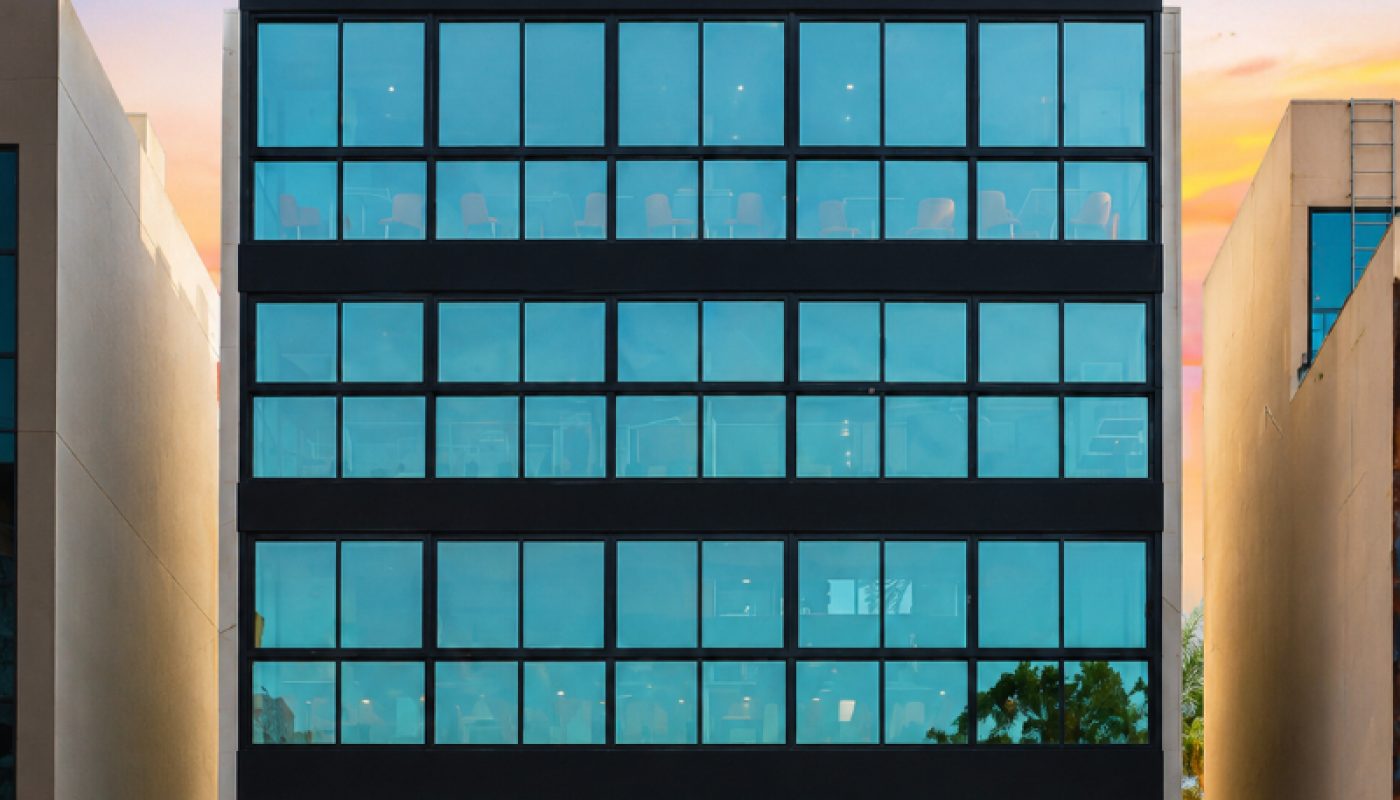 A modern, six-story glass office building is centered between two beige structures, with reflections of trees and sky visible in its windows at sunset.