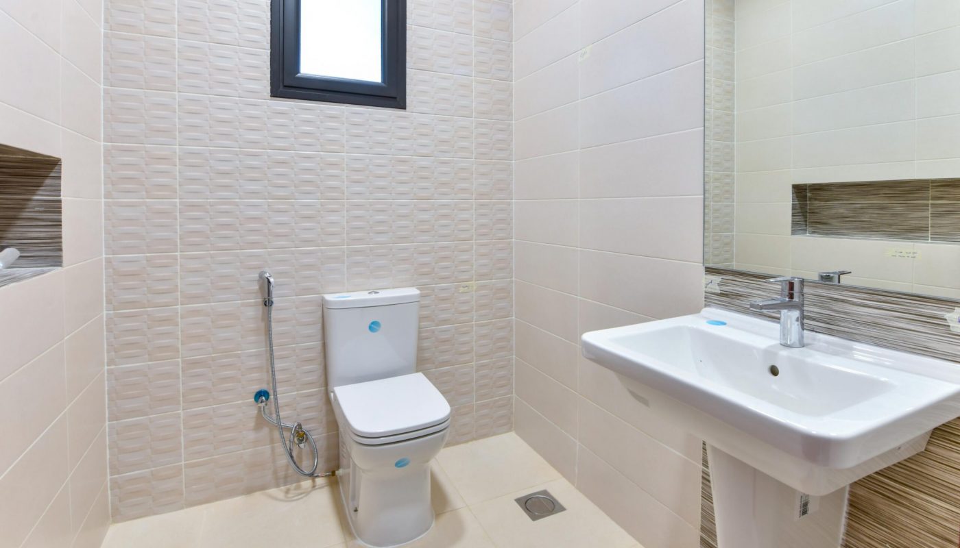 Modern bathroom with a white toilet, handheld bidet, wall-mounted sink, rectangular mirror, beige tiled walls, and a small window above the toilet.