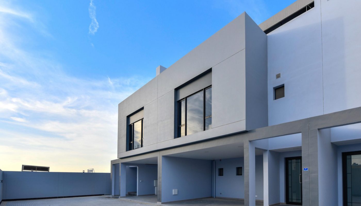 Modern two-story building with large windows, flat roof, and covered parking area, set against a blue sky with few clouds.