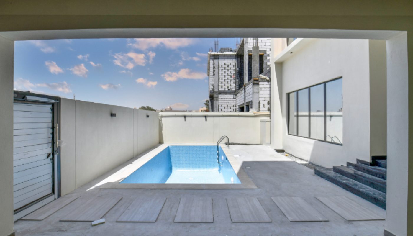 View of a small rectangular outdoor swimming pool surrounded by concrete pavement, adjacent to a white building with large windows and a closed garage door.