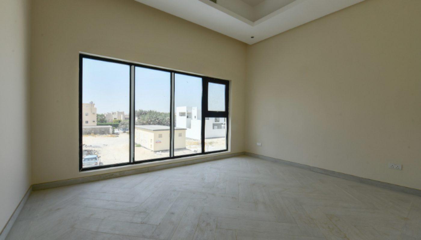 Empty room with beige walls, large floor-to-ceiling windows, tiled floor, and a view of neighboring buildings and a parking lot outside.