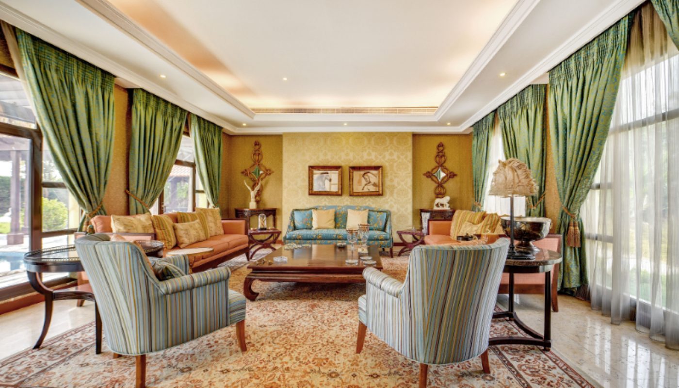 Elegant living room with patterned armchairs, a sofa, a large rug, wooden coffee tables, decorative lamps, and green curtains covering large windows.