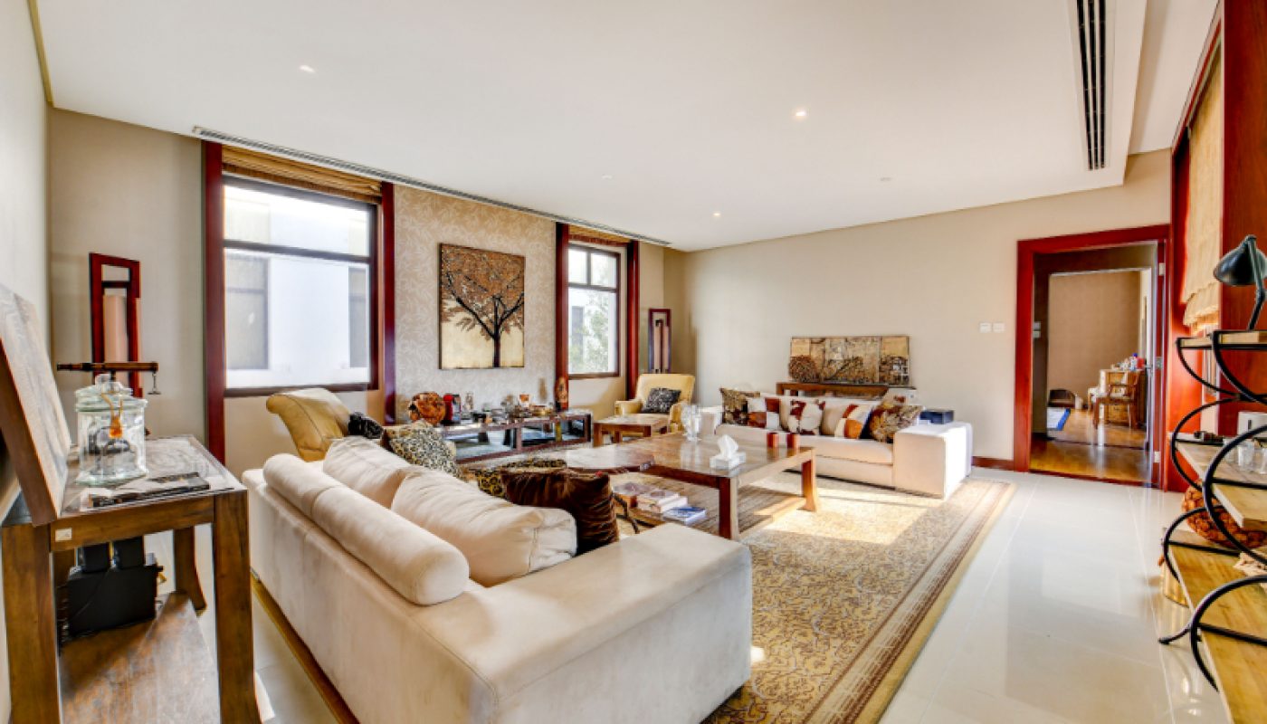 Spacious living room with cream sofas, wooden furniture, large windows, and decorative artwork on the walls. The room has neutral tones and a patterned area rug.