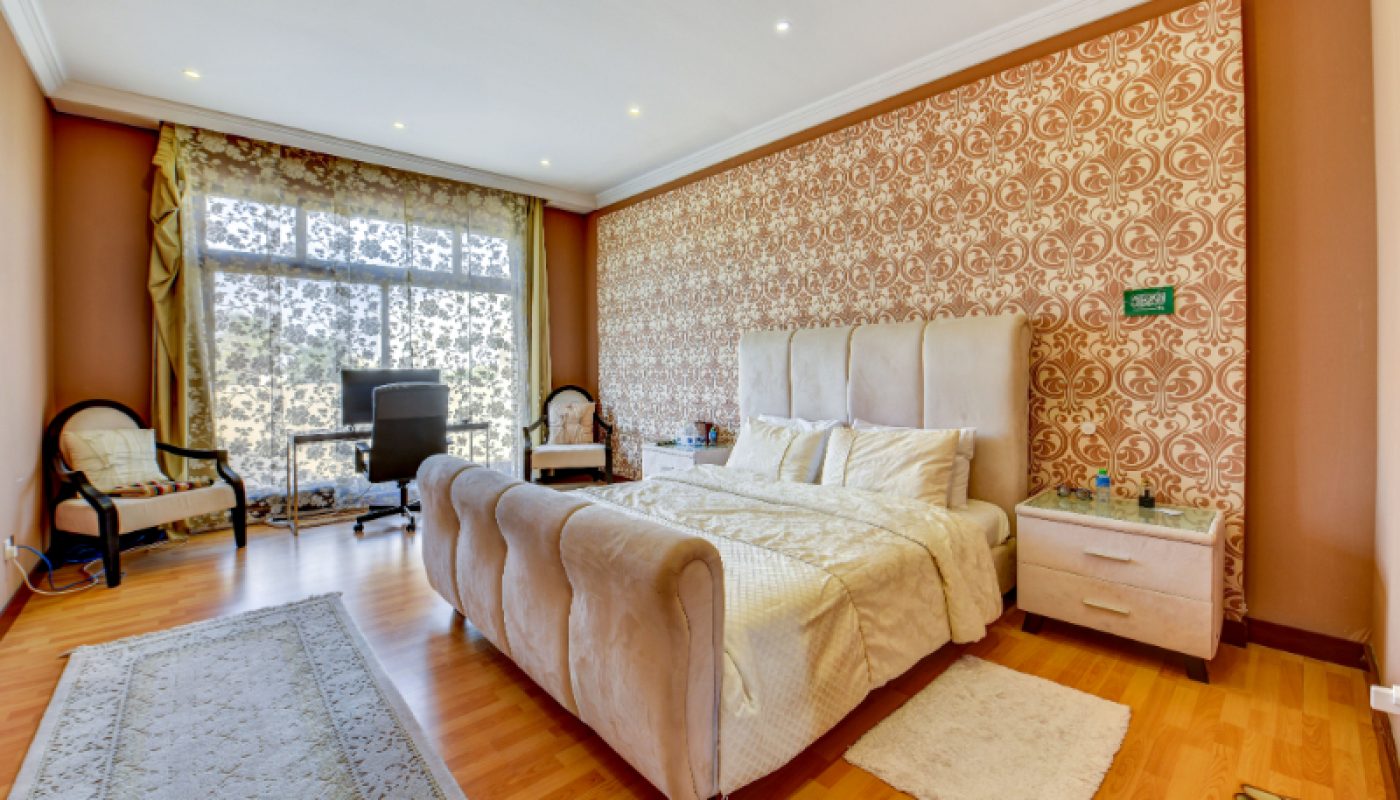 Spacious bedroom with a large bed, patterned accent wall, two nightstands, office chair at a desk, armchairs, and large windows with decorative curtains.
