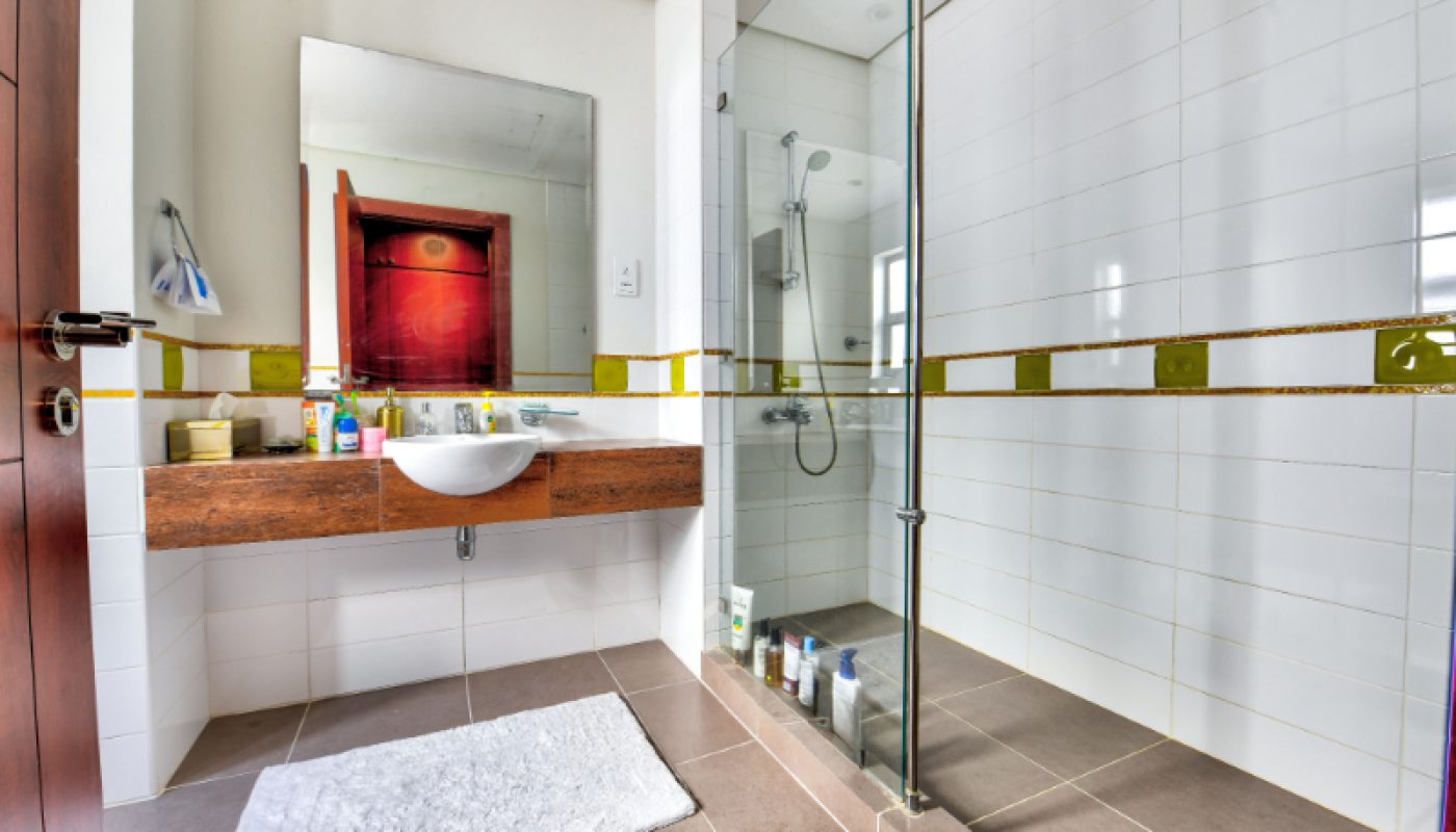 Modern bathroom with a wall-mounted sink, large mirror, toiletries, glass shower enclosure, tiled walls, and a white bath mat on the floor.