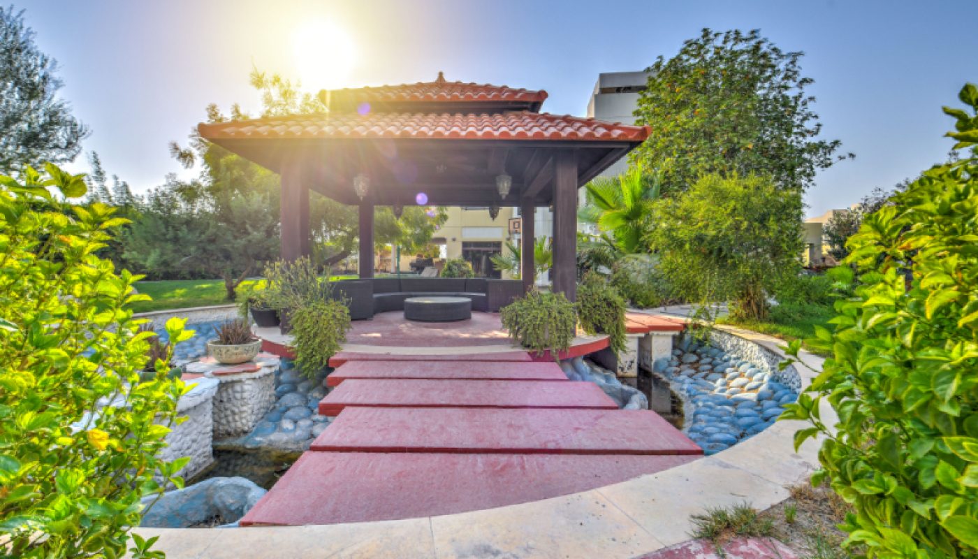 A small pavilion with a red-tiled roof sits over a stone-lined pond, surrounded by greenery, under a clear sky with the sun shining.