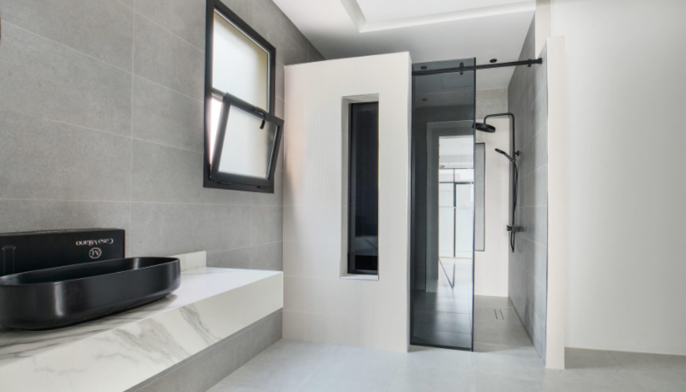 Modern bathroom with gray tiles, a black vessel sink on a white marble countertop, a glass-enclosed shower, and a frosted window.