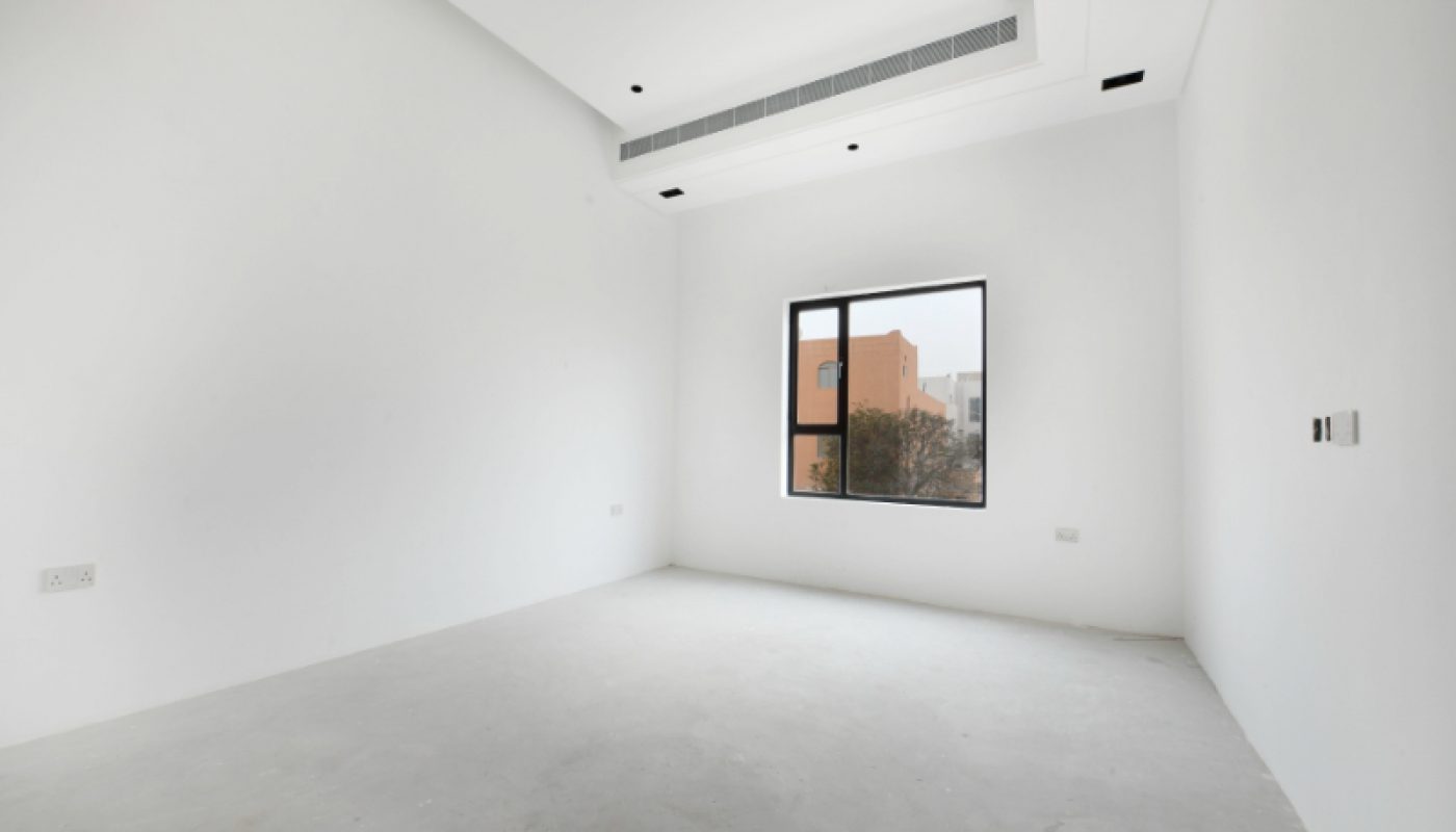 An empty, unfurnished white room with a single window, plain walls, and a ceiling vent. Natural light enters through the window, showing an outdoor building and trees.