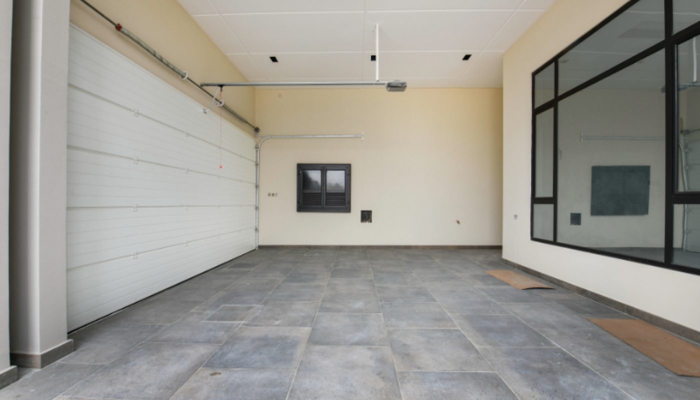 Empty modern garage with tiled floor, closed overhead door, beige walls, and large interior window. Ceiling lights and a small window are visible on the back wall.