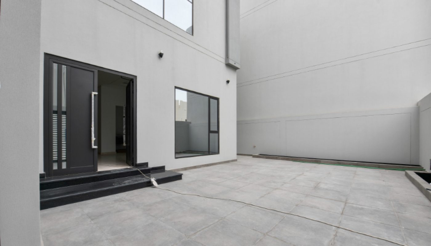 A modern, empty courtyard with gray tiles, white walls, black framed glass doors, and a single loose cable on the ground.