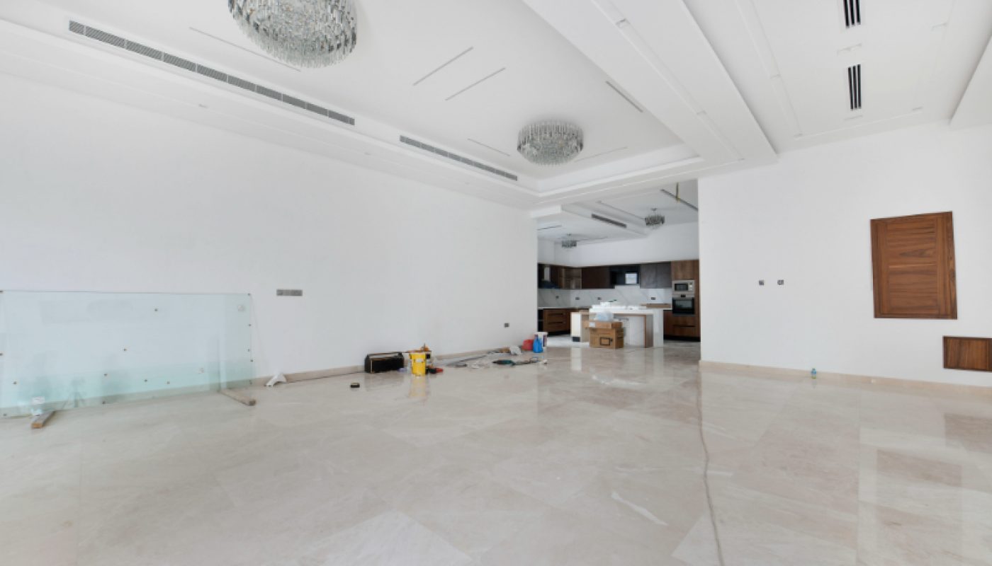 Spacious empty room with marble floors, white walls, two chandeliers, and an open kitchen area; some construction materials are scattered on the floor.