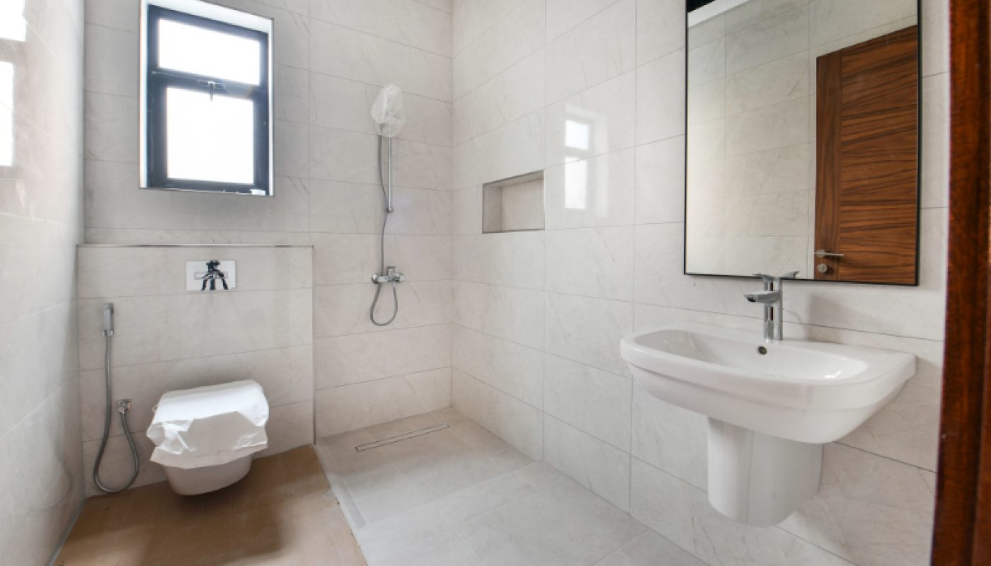Modern bathroom with white tiles, wall-mounted toilet, bidet spray, shower area, large mirror, and a wall-mounted sink. Light enters through a window on the left.