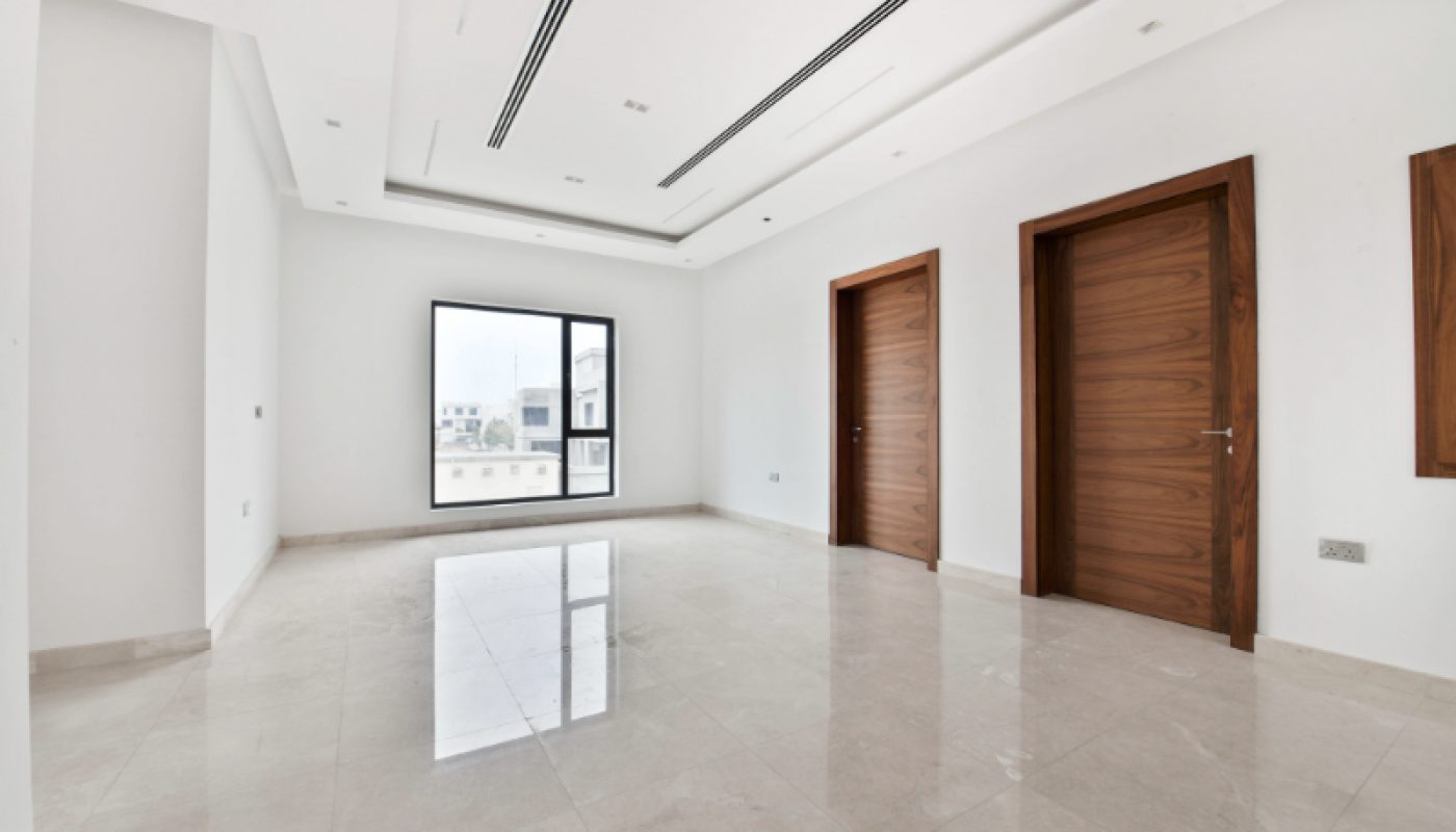 Empty, modern room with white walls, large beige tile floor, a window, and three wooden doors; recessed ceiling lighting and natural light from the window.