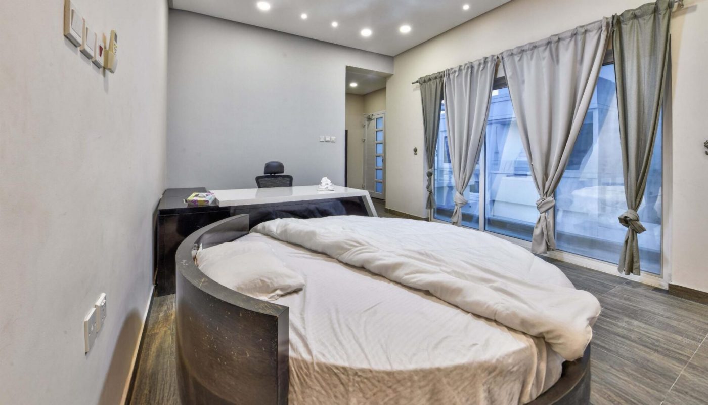A modern bedroom with a round bed, white bedding, a desk and chair, large windows with curtains, and wood flooring.