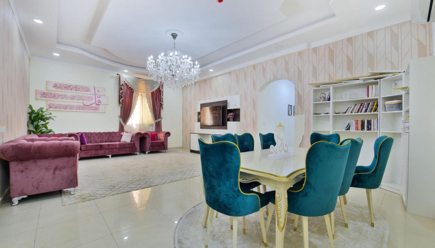 A modern living and dining area with teal chairs, a white table, purple sofas, a chandelier, a wall-mounted TV, bookshelves, and neutral decor.