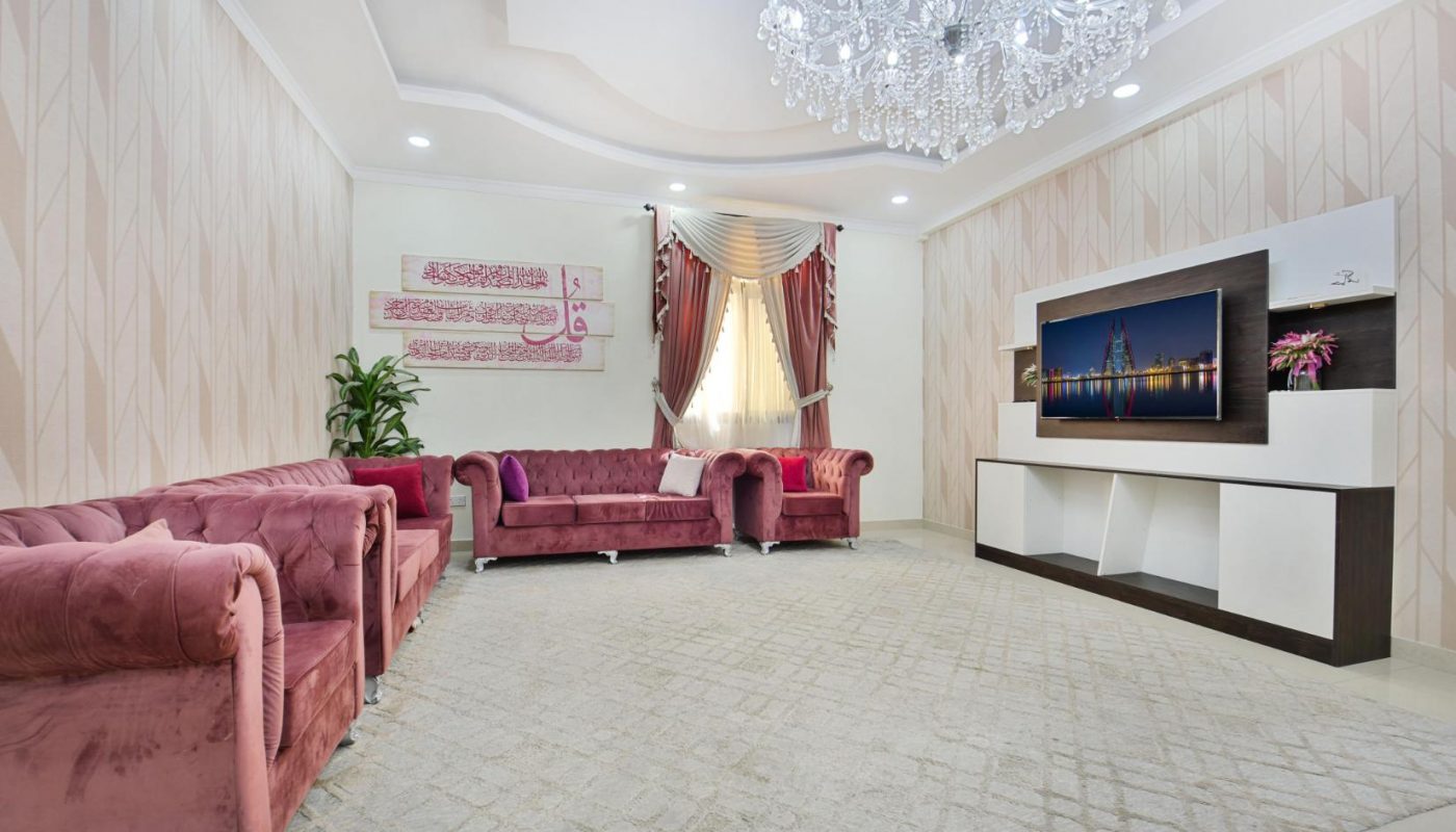 A modern living room with pink velvet sofas, a chandelier, patterned wallpaper, a TV on a white entertainment unit, and large windows with curtains.