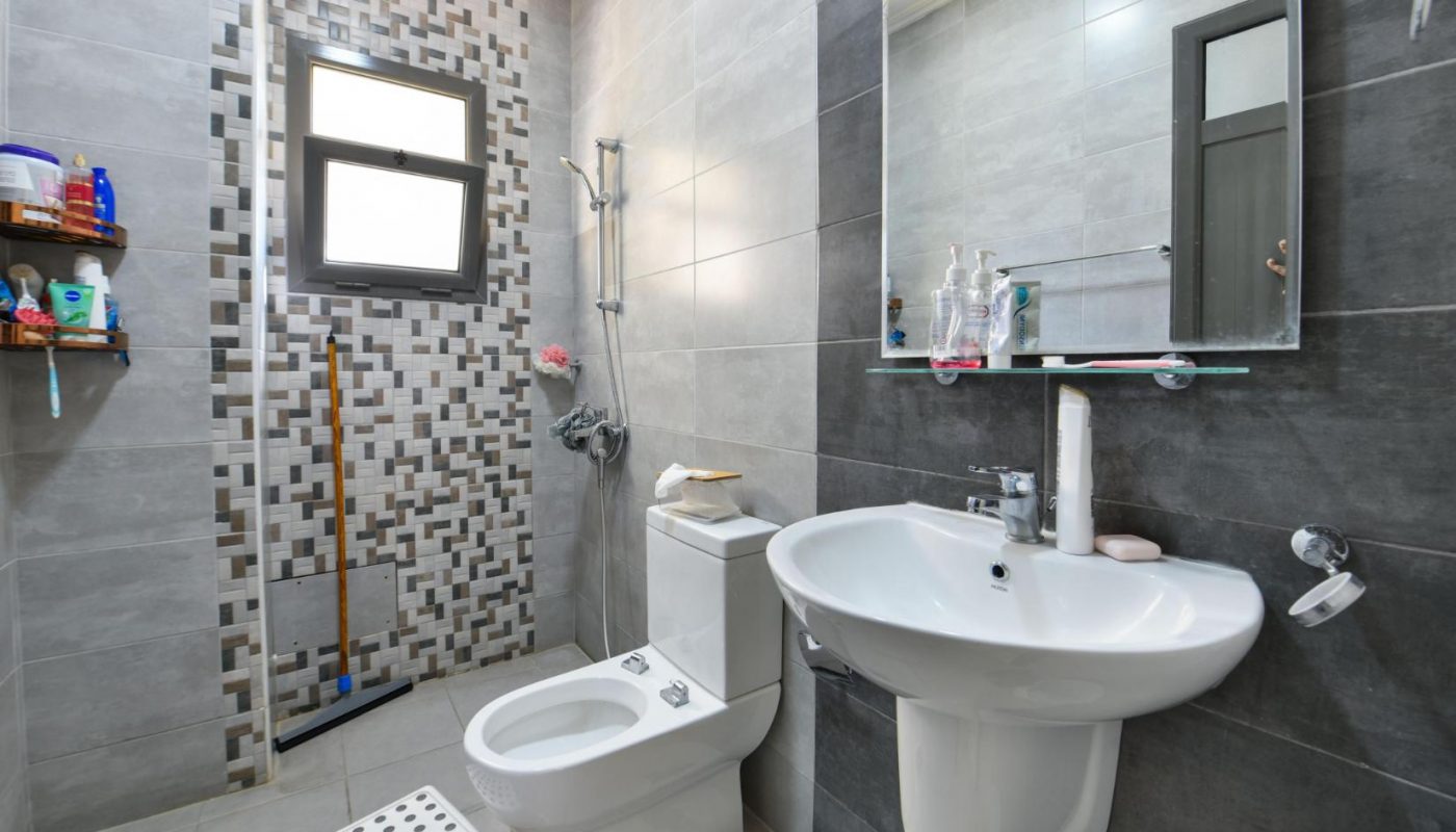 Modern bathroom with gray tiled walls, a sink, a wall-mounted mirror, a toilet, a bidet, a tiled shower area, shelves with toiletries, and a small white step stool.