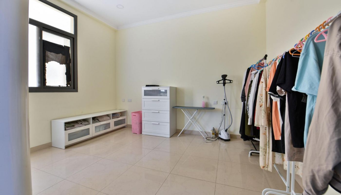 A tidy room with tiled floor, a clothes rack, dresser, ironing board, laundry basket, and storage bench under a large window.