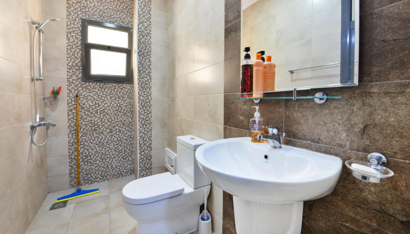 Modern bathroom with beige tiles, wall-mounted sink, toilet, small window, walk-in shower, and cleaning supplies including a mop and broom. Toiletries are placed on a glass shelf above the sink.