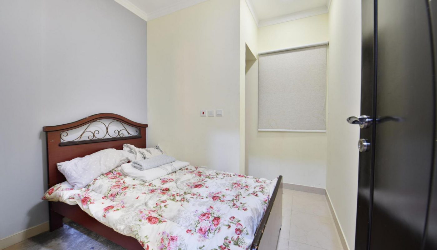 A small bedroom with a wooden bed covered in floral bedding, pillows, and towels, beside cream-colored walls and a window with a closed blind.