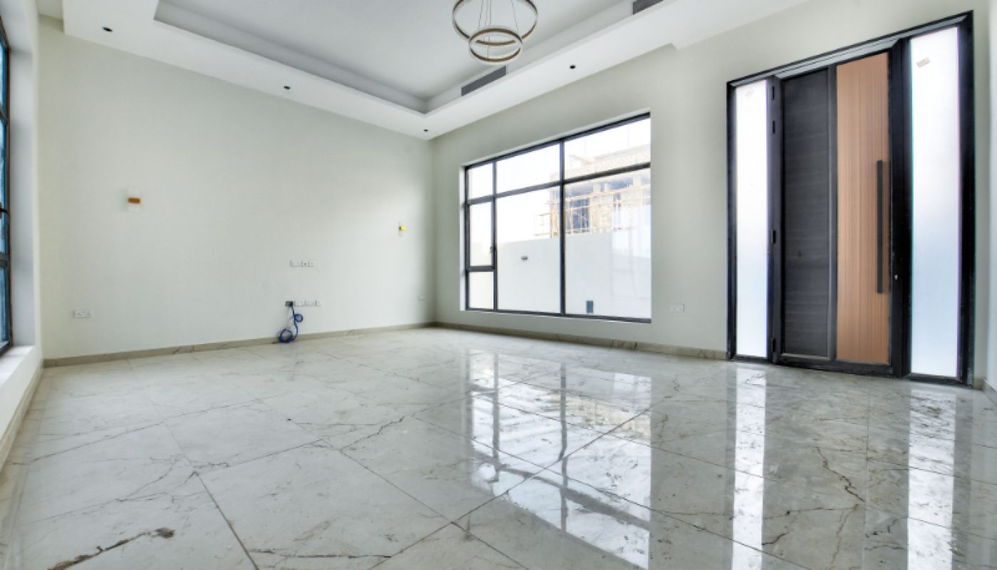 A spacious, empty room with marble floors, large windows, an open door, white walls, and a small scooter near the corner.
