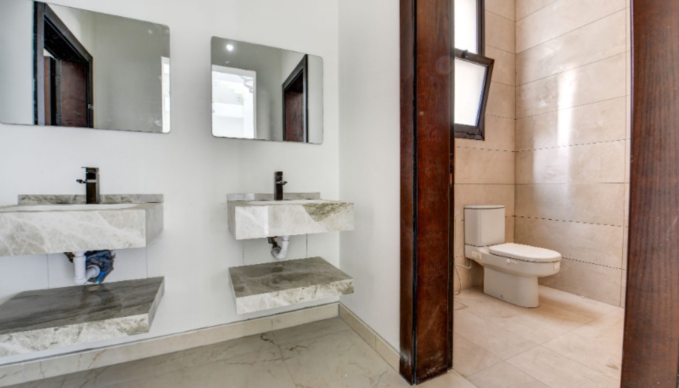 Modern bathroom with two wall-mounted sinks and mirrors on the left, separated by a partial wall from a toilet and window on the right. Stone countertops and tiled floors throughout.