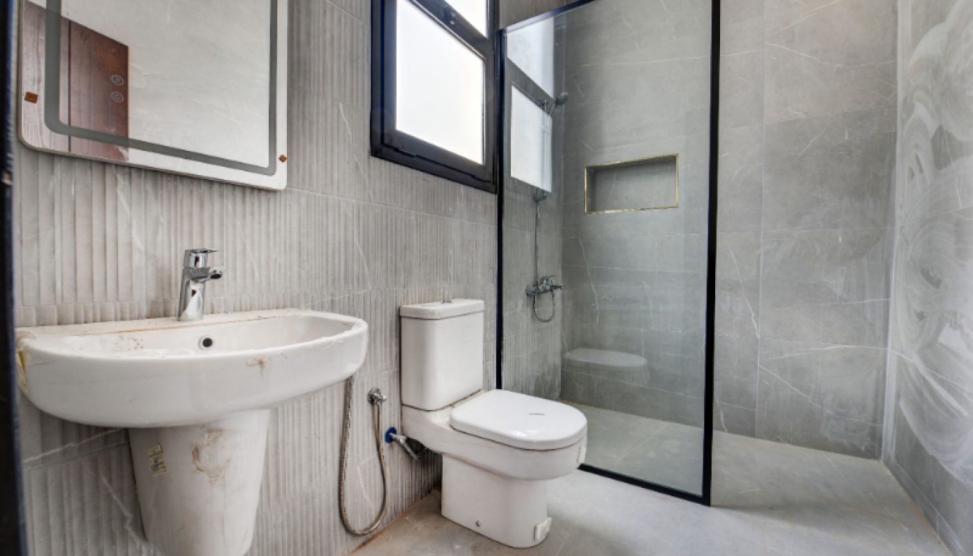 Modern bathroom with a wall-mounted sink, toilet, and glass-enclosed shower; gray tiled walls and floor, window above sink and toilet.
