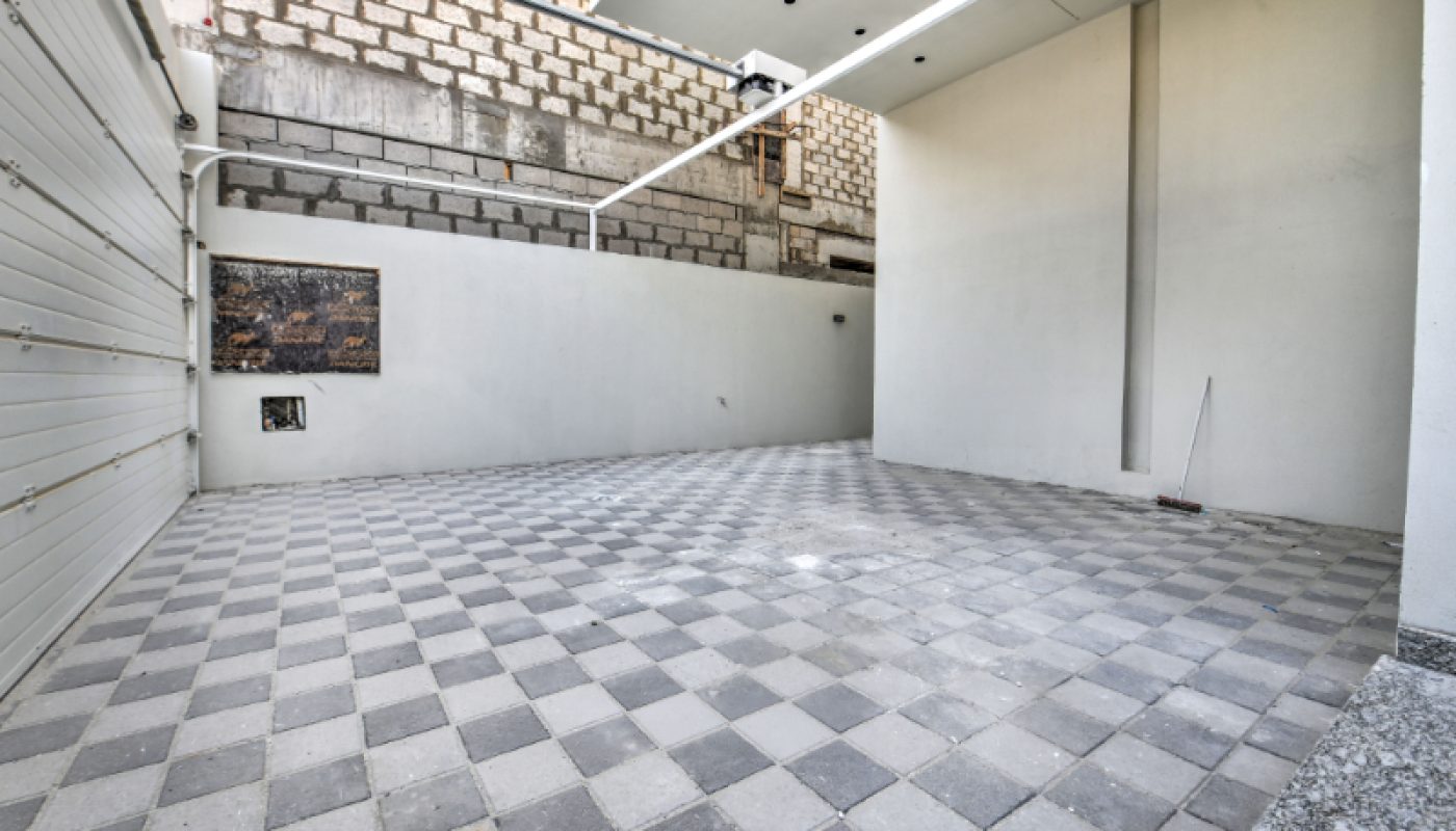 Empty covered garage with a checkered tile floor, white walls, and a partially unfinished brick wall in the background. A broom is leaning against the right wall.