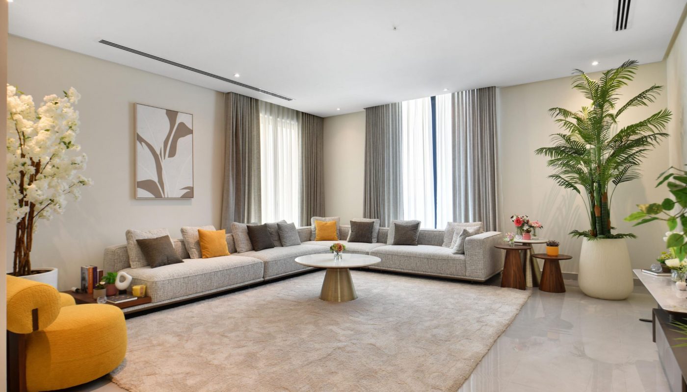 Modern living room with a large L-shaped sofa, neutral decor, a round coffee table, patterned rug, and several potted plants, illuminated by natural light from tall windows.