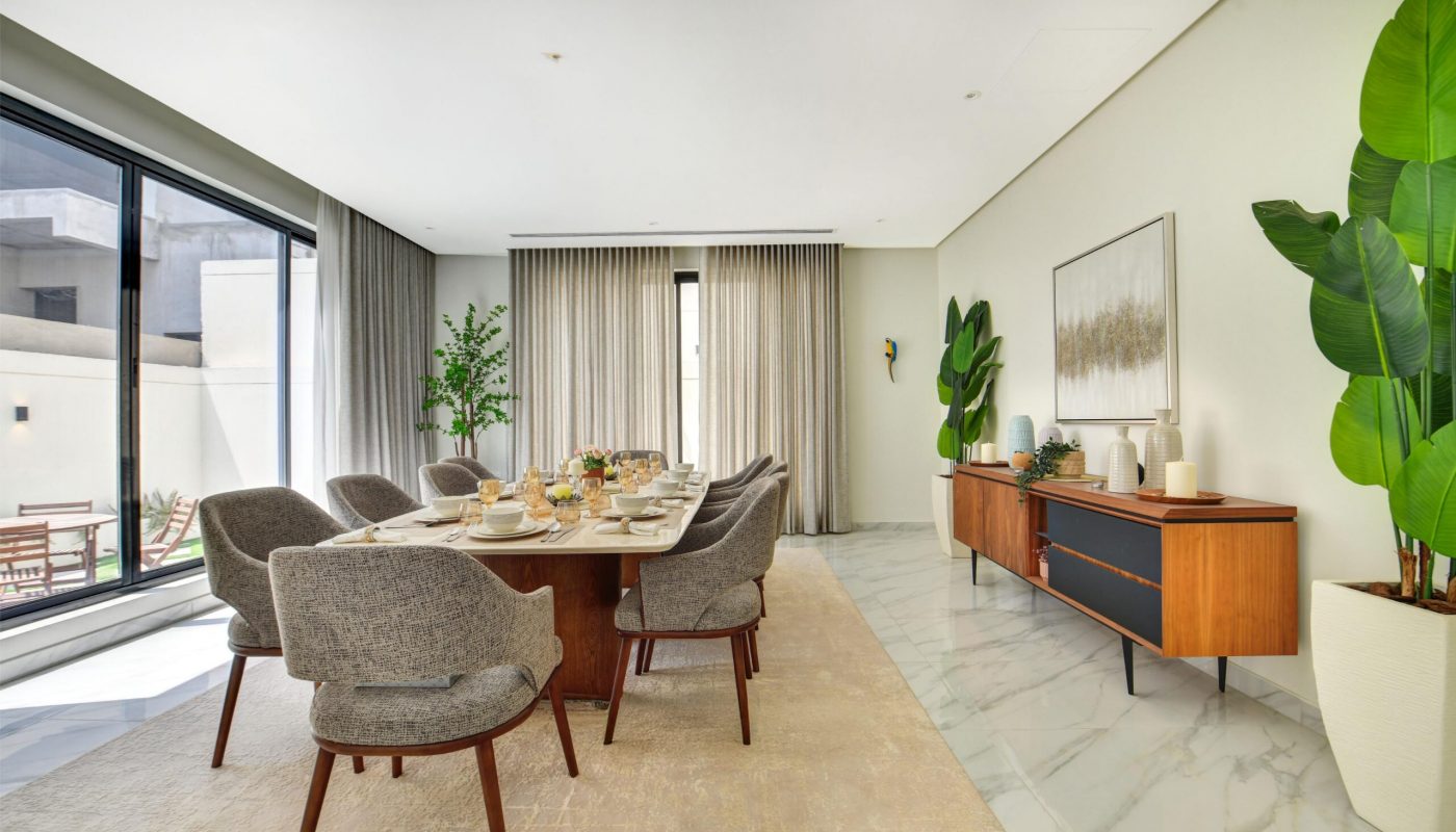 Modern dining room with a wooden table set for six, gray upholstered chairs, sideboard, potted plants, large window, and neutral decor.