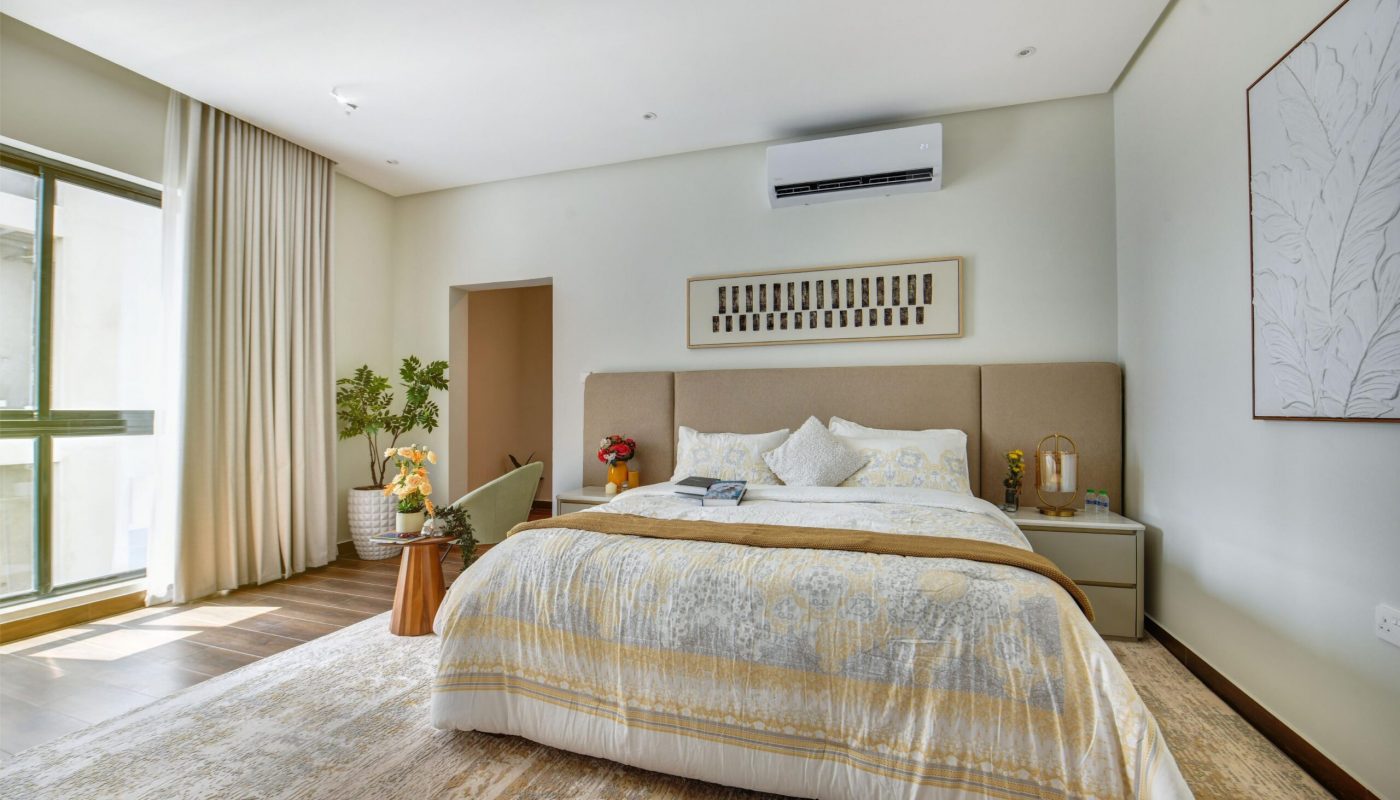 A modern bedroom with a large bed, light-colored bedding, side tables, potted plants, wall art, and an air conditioner above the headboard. Natural light enters through a window with curtains.