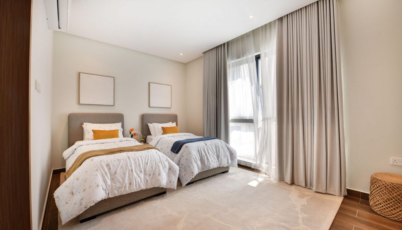 Modern bedroom with two single beds, a large window with sheer curtains, minimalist wall art, and a woven ottoman on a light rug over wooden flooring.