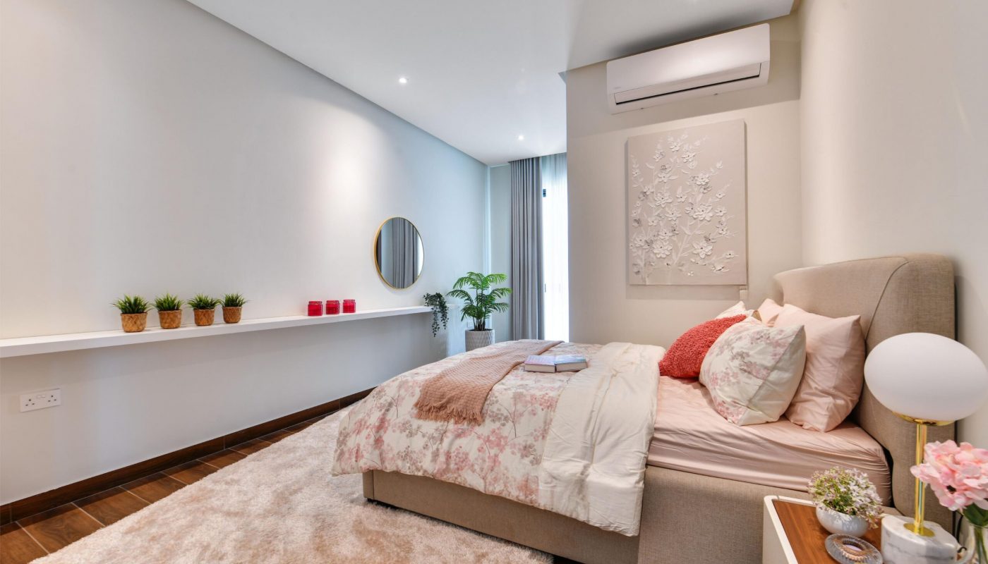 Modern bedroom with a beige bed, pink and white bedding, a round mirror, potted plants on a shelf, a white rug, and a wall-mounted air conditioner.