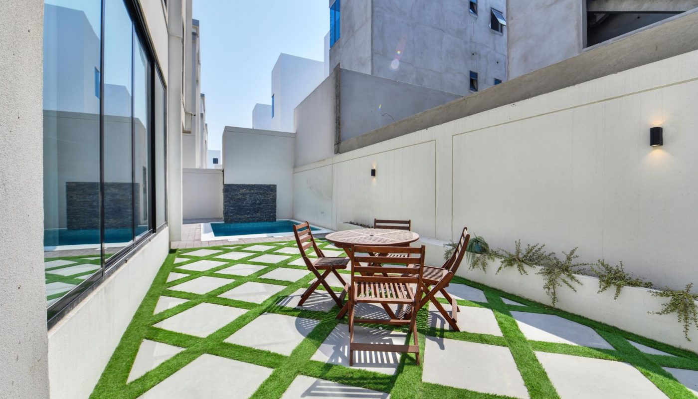 Outdoor patio with a round wooden table and four chairs on a geometric stone and grass floor, next to a white wall and a swimming pool in the background.