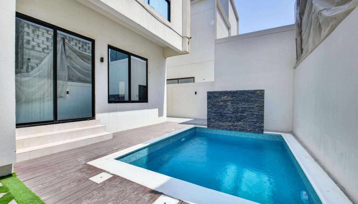 A small modern backyard pool with a stone water feature, wooden deck, and white surrounding walls next to a house with large glass doors.