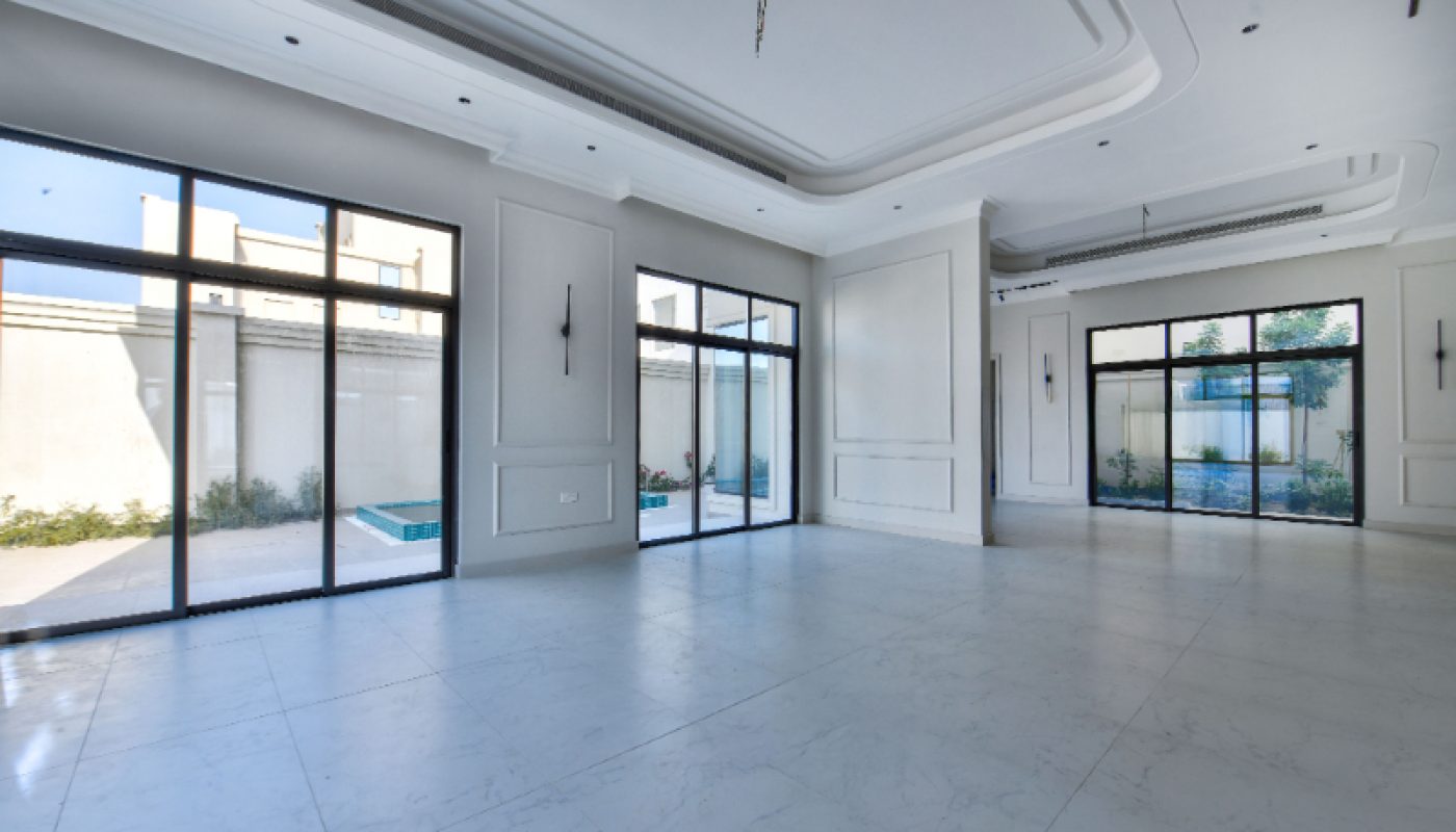 Spacious, empty modern room with large floor-to-ceiling windows, white marble flooring, recessed ceiling, and views of a small outdoor pool and garden.
