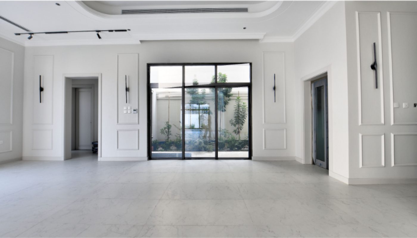 A modern, empty room with white walls and marble flooring, large glass doors leading to a garden, and minimal black lighting fixtures.