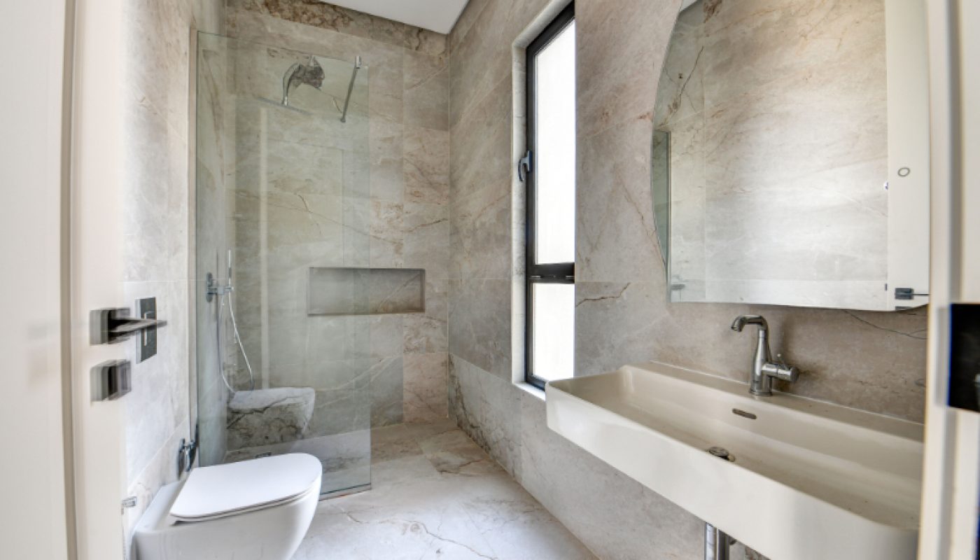 Modern bathroom with beige marble walls and floor, wall-mounted toilet, glass-enclosed shower, large window, and rectangular sink with mirror above.