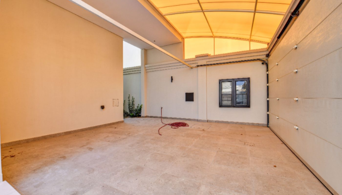 Covered garage space with a light-colored tiled floor, beige walls, a window, and a rolled-up hose on the ground. The ceiling is topped with a translucent yellow canopy.