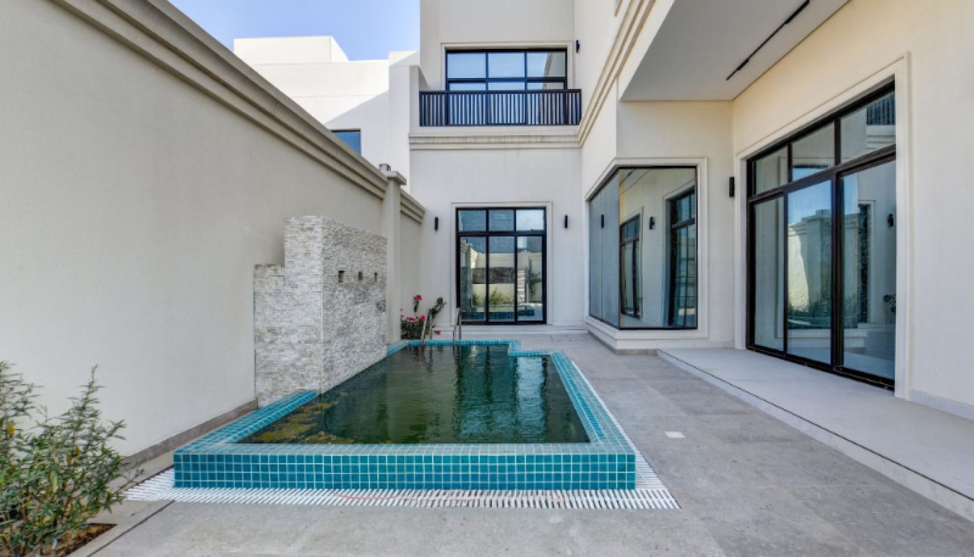 Rectangular plunge pool with tiled edges next to a modern beige building, large windows, and a stone wall water feature.