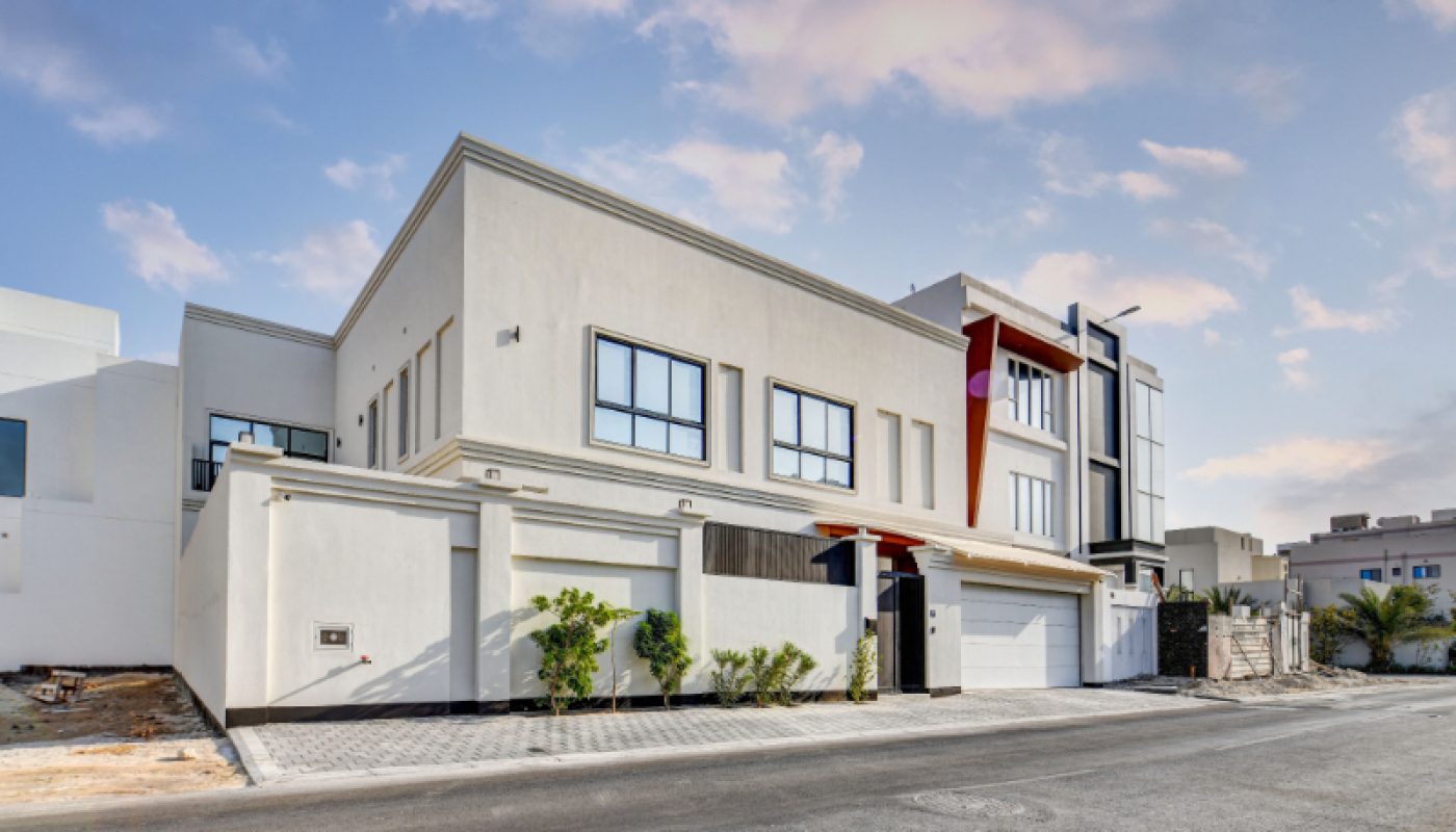 Modern two-story white house with large windows, a garage, and minimal landscaping, situated on a paved street under a partly cloudy sky.