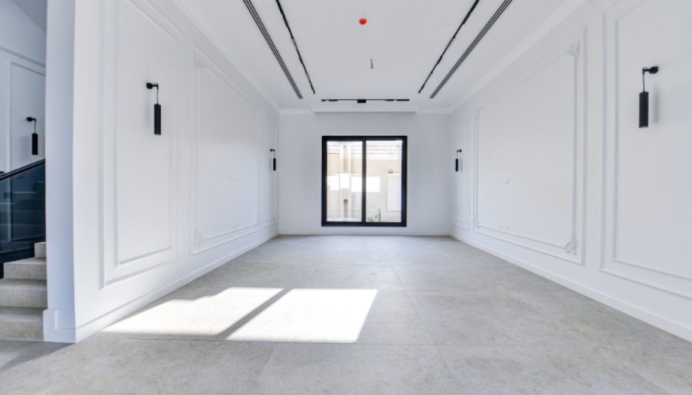 Spacious, empty white room with tile flooring, modern wall sconces, a large window, and a staircase partially visible on the left.