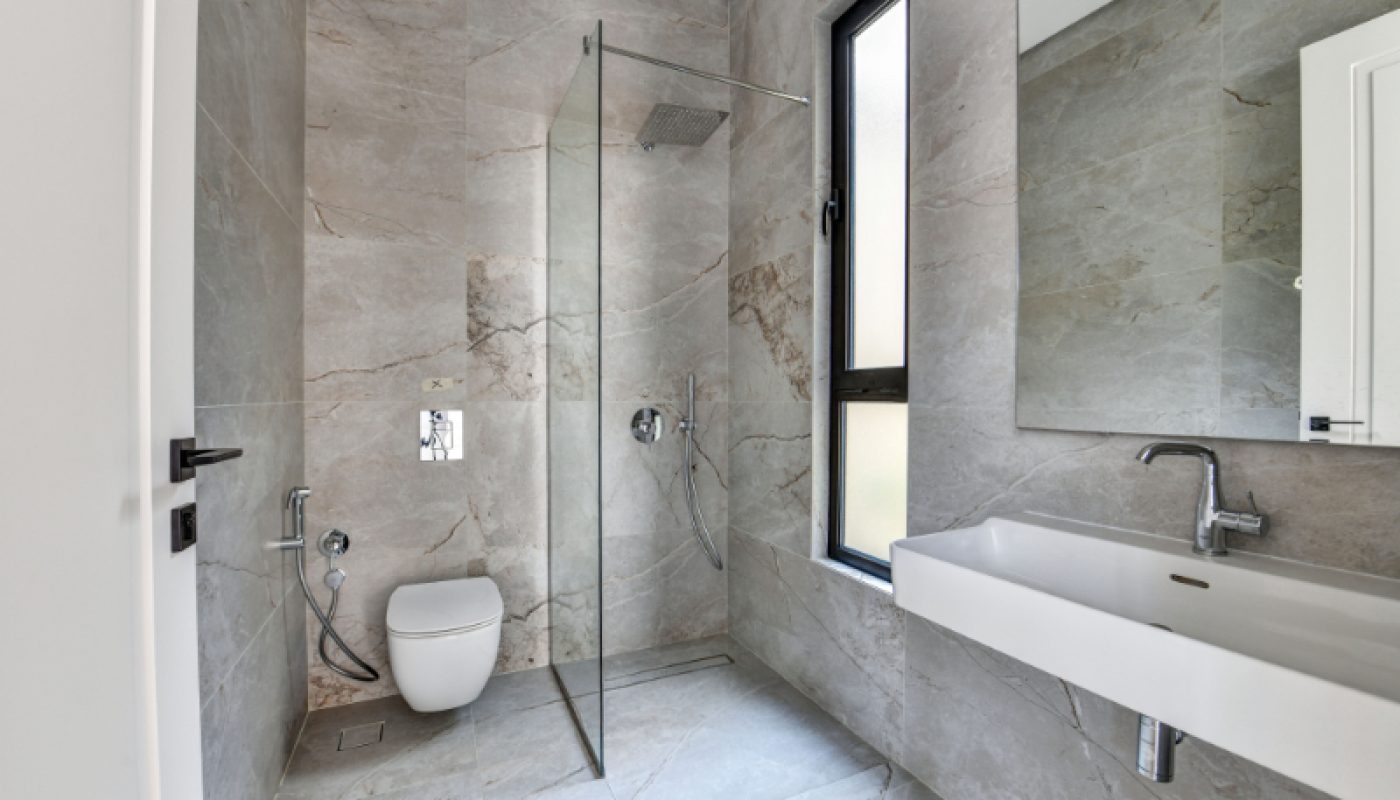 Modern bathroom with gray marble walls, glass-enclosed shower, wall-mounted toilet, large rectangular sink, and a window allowing natural light.