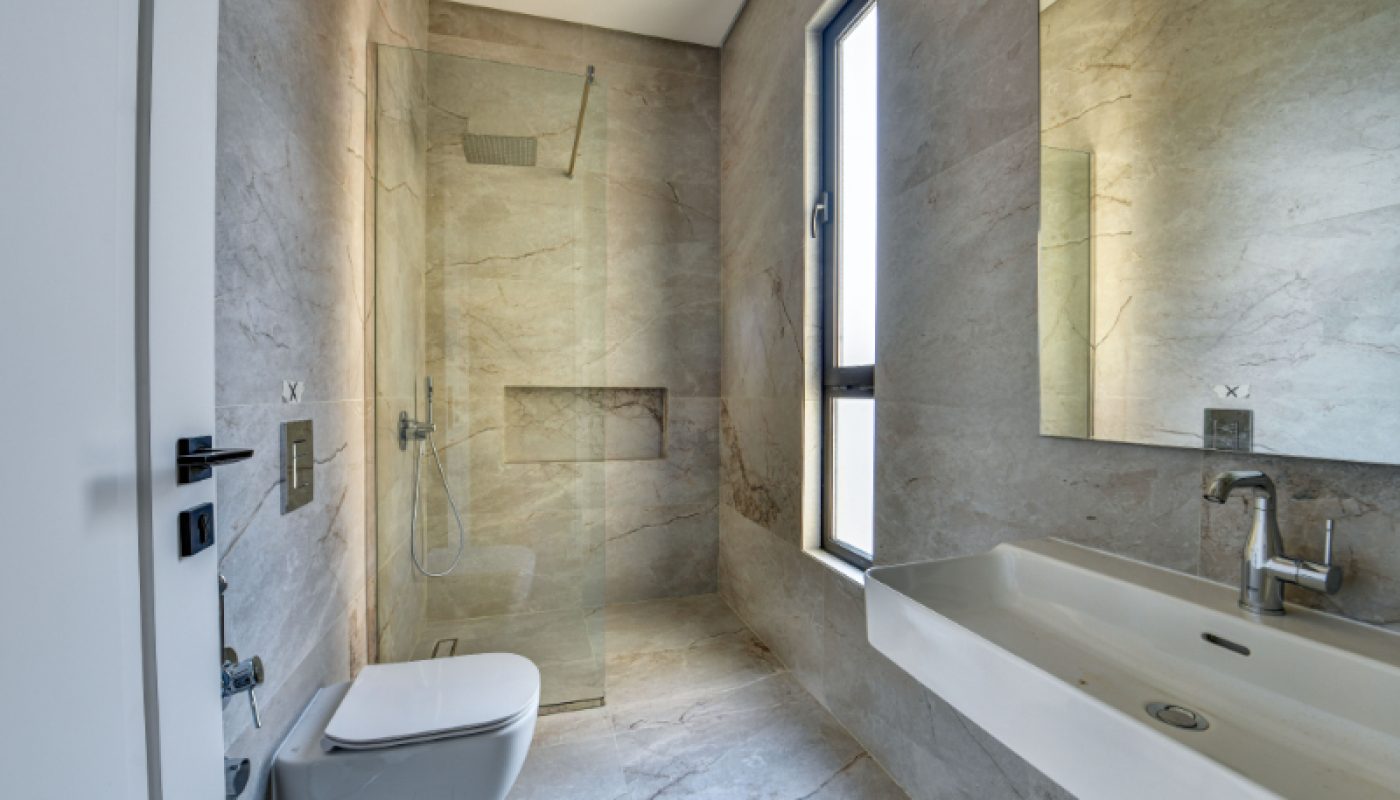 Modern bathroom with a wall-mounted toilet, walk-in glass shower, large rectangular sink, and a window; all surfaces feature light beige marble tiles.