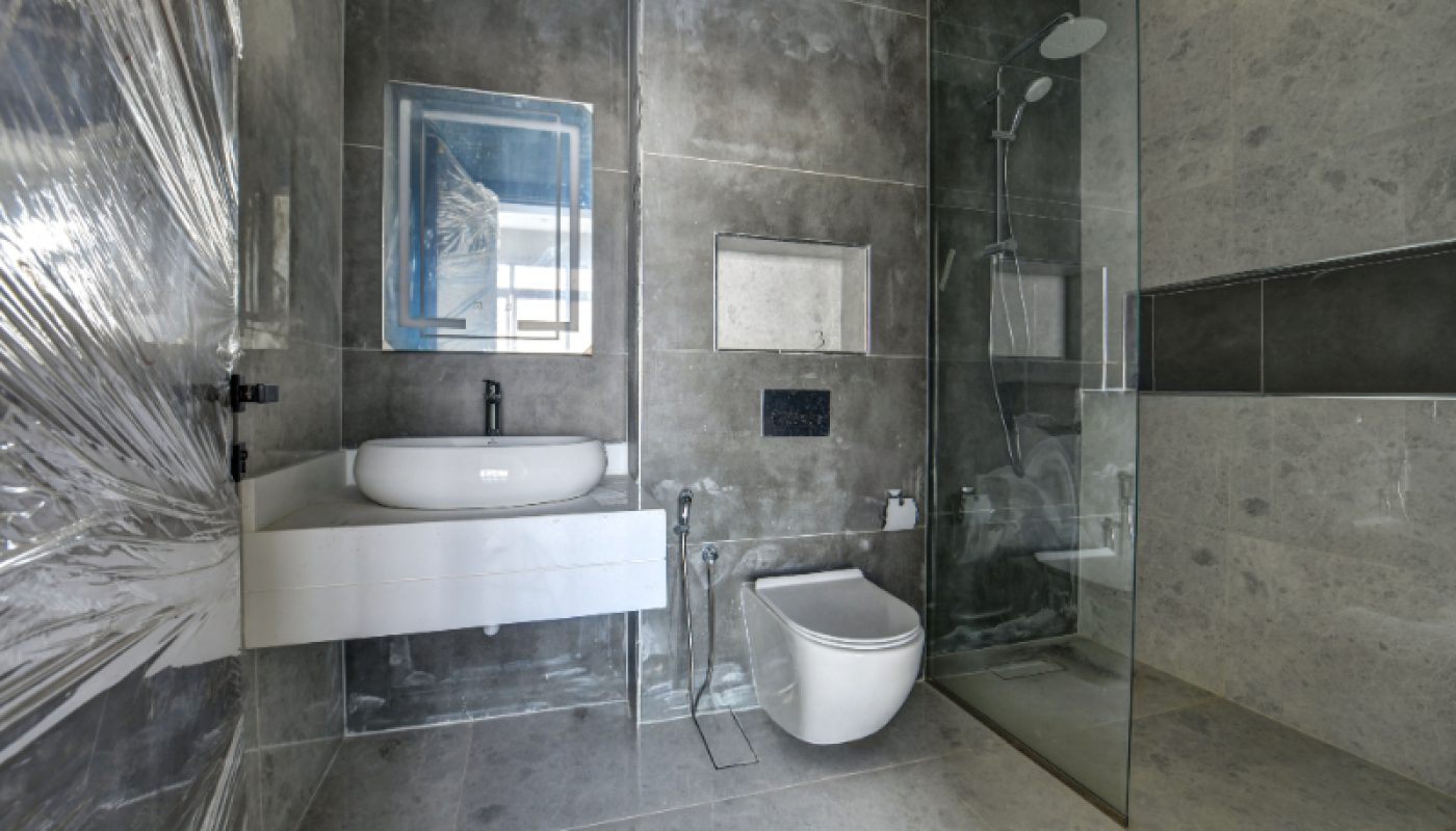 Modern bathroom with gray tiles, a wall-mounted white sink, a mirror, a wall-hung toilet, and a glass-enclosed shower area. Plastic wrap covers part of the left wall.