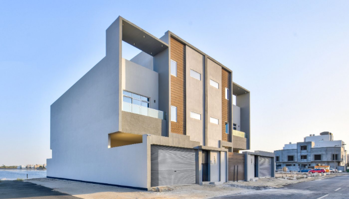 Modern three-story residential building with large windows, balconies, and three garage doors, situated on a paved street with another building visible in the background.