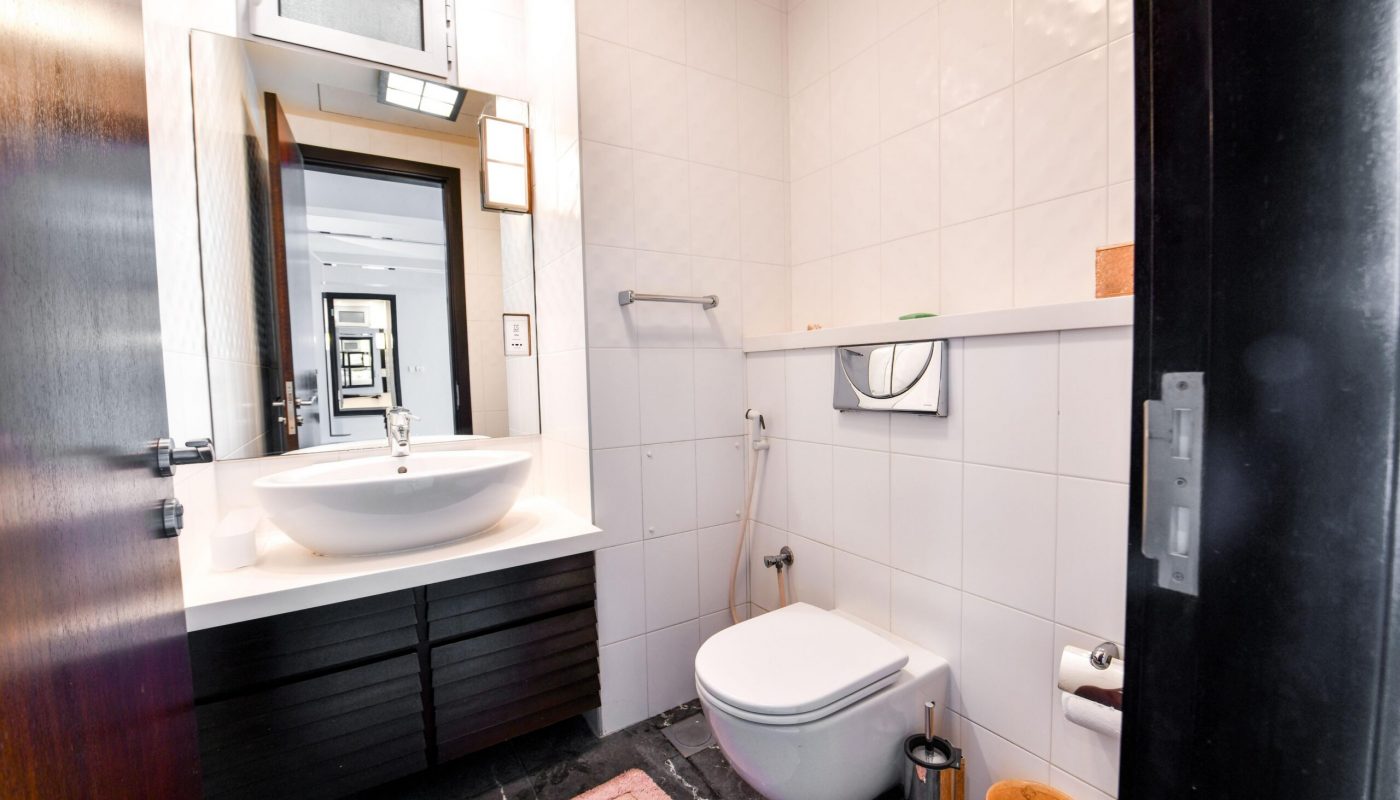 Modern bathroom with a white sink, wall-mounted toilet, bidet spray, mirror, towel rail, and a pink bath mat on a dark tiled floor.
