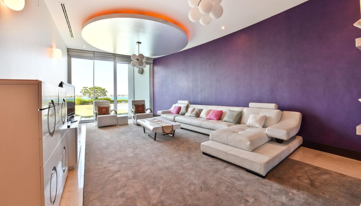 Modern living room with a purple accent wall, curved white sectional sofa with colorful pillows, large windows, and contemporary lighting fixtures.