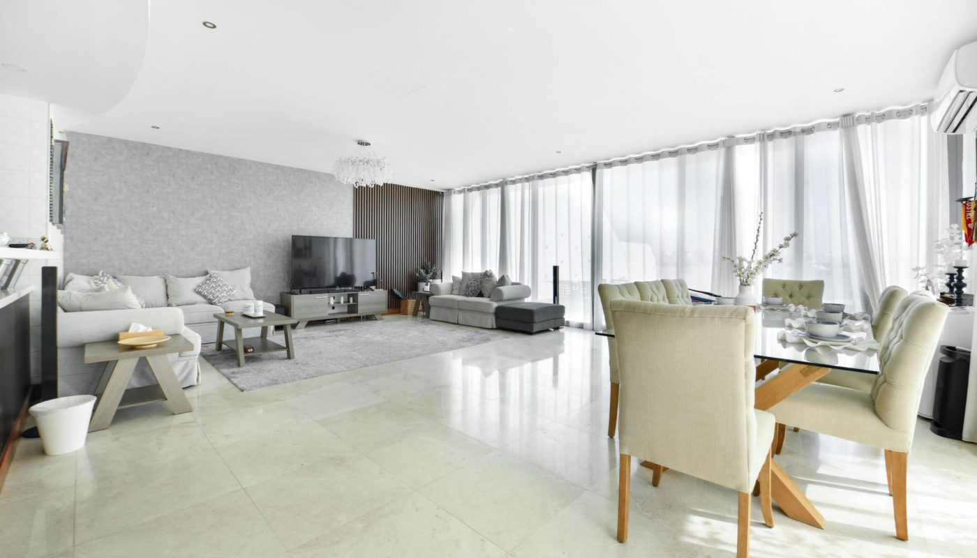 Spacious living and dining area with light flooring and walls. Features a large sectional sofa, TV, coffee tables, dining set, and sheer drapes covering floor-to-ceiling windows.